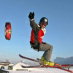 Ein Skifahrer mit Helm und Skibrille springt auf einer verschneiten Piste und hält dabei einen Griff, der an einem Drachen im Hintergrund befestigt ist.