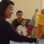 Eine Frau hält zwei Handpuppen vor einer Gruppe von sitzenden Erwachsenen und Kindern in einem Klassenzimmer hoch.