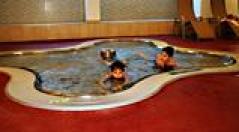 Drei Kinder schwimmen in einem kleinen, unregelmäßig geformten Hallenbad, das von einem roten Bodenbelag umgeben ist.