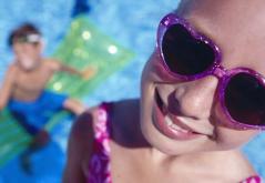 Nahaufnahme eines Kindes mit herzförmiger Sonnenbrille, das in die Kamera lächelt, mit einem anderen Kind auf einem grünen Schwimmer in einem Schwimmbad im Hintergrund.