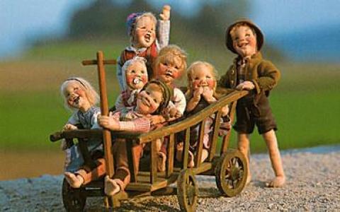 Sieben lächelnde Puppen in altmodischer Kleidung, sechs davon in einem Holzkarren sitzend und eine daneben stehend, vor einem unscharfen Hintergrund im Freien.