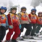 Eine Gruppe von Kindern in bunten Skiklamotten und mit Helmen steht in einer Reihe auf einer verschneiten Piste, begleitet von einem erwachsenen Skilehrer.