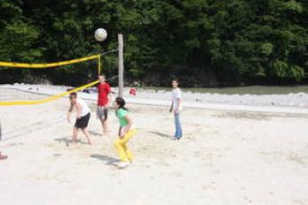 Vier Personen spielen Beachvolleyball auf einem Sandplatz, im Hintergrund sind Bäume zu sehen.