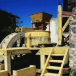 Hölzerne Wassermühle mit Wasserrad, Steinmauer und Stufen, die zum Mahlen von Getreide verwendet wird, im Freien unter blauem Himmel.