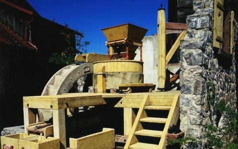Hölzerne Wassermühle mit Wasserrad, Steinmauer und Stufen, die zum Mahlen von Getreide verwendet wird, im Freien unter blauem Himmel.