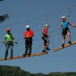 Fünf Personen mit Helmen und Klettergurten überqueren eine Hängeseilbrücke im Freien, mit Bäumen und Bergen im Hintergrund.