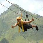 Eine Person, die einen Helm und ein Gurtzeug trägt, fährt auf einer Seilrutsche in einem Gebirge und zeigt mit beiden Händen den Daumen nach oben.