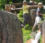 Drei Kinder stehen in der Nähe eines hölzernen Wasserkanals zwischen großen Felsen, durch den Wasser fließt, während im Hintergrund Grünpflanzen zu sehen sind.
