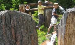 Drei Kinder stehen in der Nähe eines hölzernen Wasserkanals zwischen großen Felsen, durch den Wasser fließt, während im Hintergrund Grünpflanzen zu sehen sind.