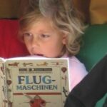 Ein Kind mit blondem Haar liest aufmerksam ein deutsches Buch mit dem Titel "Flugmaschinen", auf dessen Umschlag eine Flugmaschine abgebildet ist.
