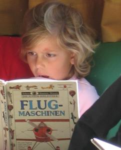 Ein Kind mit blondem Haar liest aufmerksam ein deutsches Buch mit dem Titel "Flugmaschinen", auf dessen Umschlag eine Flugmaschine abgebildet ist.