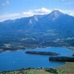 Ein großer blauer See mit einer Insel in der Mitte, umgeben von grünen Feldern und Wäldern, mit Bergen im Hintergrund unter einem teilweise bewölkten Himmel.
