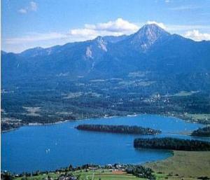 Ein großer blauer See mit einer Insel in der Mitte, umgeben von grünen Feldern und Wäldern, mit Bergen im Hintergrund unter einem teilweise bewölkten Himmel.