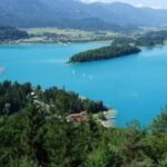 Luftaufnahme eines türkisfarbenen Sees, umgeben von grünen Wäldern, Inseln und entfernten Bergen unter einem teilweise bewölkten Himmel.