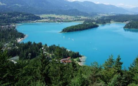Luftaufnahme eines türkisfarbenen Sees, umgeben von grünen Wäldern, Inseln und entfernten Bergen unter einem teilweise bewölkten Himmel.