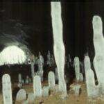 Mehrere hohe, vertikale Eisformationen ragen aus dem Höhlenboden, und im Hintergrund ist ein beleuchteter Höhleneingang zu sehen.