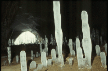 Mehrere hohe, vertikale Eisformationen ragen aus dem Höhlenboden, und im Hintergrund ist ein beleuchteter Höhleneingang zu sehen.