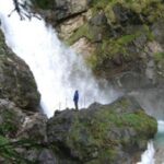 Eine Person in einer blauen Jacke steht auf einem Felsvorsprung in der Nähe eines Wasserfalls, umgeben von moosbewachsenen Felsen und Grünpflanzen.