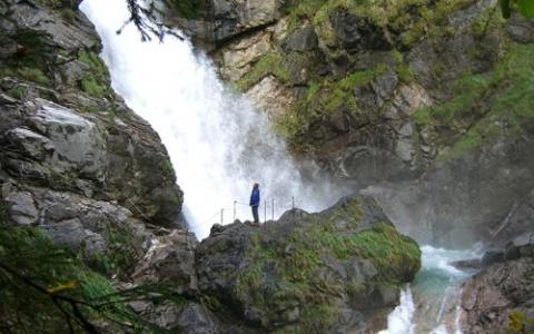 Eine Person in einer blauen Jacke steht auf einem Felsvorsprung in der Nähe eines Wasserfalls, umgeben von moosbewachsenen Felsen und Grünpflanzen.