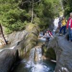 Vier Personen stehen und kauern auf Felsen neben einem kleinen, in Kaskaden fließenden Bach in einem Waldgebiet, umgeben von Bäumen und Grünzeug.