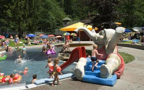 Kinder spielen in einem flachen Außenbecken mit einer roten Rutsche in Form eines Elefanten; Erwachsene und Kinder entspannen sich auf dem Rasen und den Liegestühlen in der Nähe.