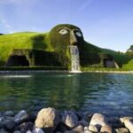 Große Rasenskulptur in Form eines Kopfes, aus dessen Mund Wasser in einen Teich fließt, in einem begrünten Außenbereich.