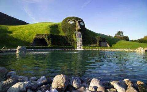 Große Rasenskulptur in Form eines Kopfes, aus dessen Mund Wasser in einen Teich fließt, in einem begrünten Außenbereich.