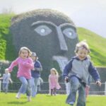 Eine Gruppe von Kindern rennt über eine Wiese vor einer großen Heckenskulptur in Form eines Gesichts, aus dessen Mund Wasser fließt.