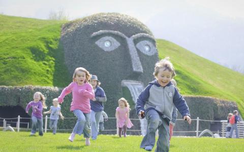 Eine Gruppe von Kindern rennt über eine Wiese vor einer großen Heckenskulptur in Form eines Gesichts, aus dessen Mund Wasser fließt.