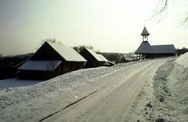 Schneebedeckte Straße, die zu einer Ansammlung von Gebäuden mit steilen Dächern und einem kleinen Glockenturm führt, umgeben von Schnee in einer ländlichen Winterlandschaft.