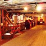 Das Innere eines alten Holzgebäudes mit Balken, historischen Maschinen und Geräten an den Wänden, beleuchtet von warmen Deckenlampen.