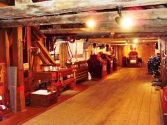 Das Innere eines alten Holzgebäudes mit Balken, historischen Maschinen und Geräten an den Wänden, beleuchtet von warmen Deckenlampen.