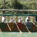 Eine Gruppe von Personen mit Wikingerhelmen rudert ein Holzboot mit bunten Schilden an der Seite auf einem Fluss entlang, geführt von einer Person, die hinten steht.