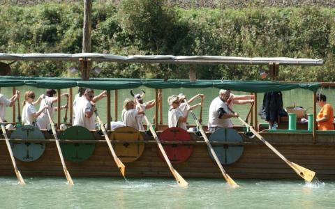 Eine Gruppe von Personen mit Wikingerhelmen rudert ein Holzboot mit bunten Schilden an der Seite auf einem Fluss entlang, geführt von einer Person, die hinten steht.