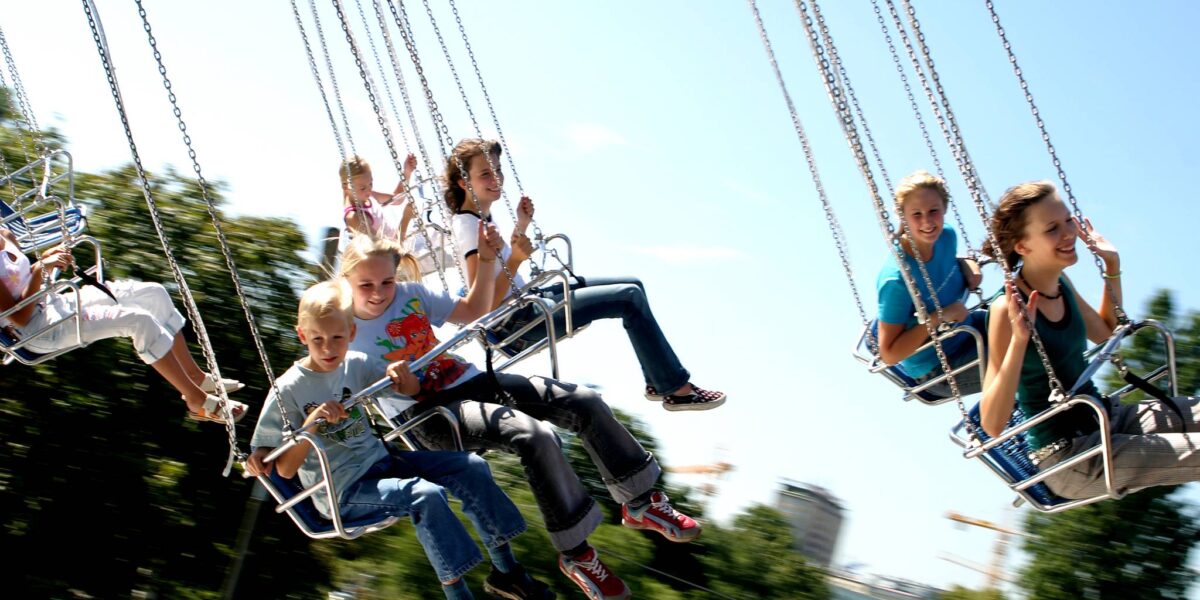 Kinder fahren auf einem sich drehenden Schaukelkarussell in einem Vergnügungspark unter einem klaren blauen Himmel, mit Bäumen und Gebäuden im Hintergrund.