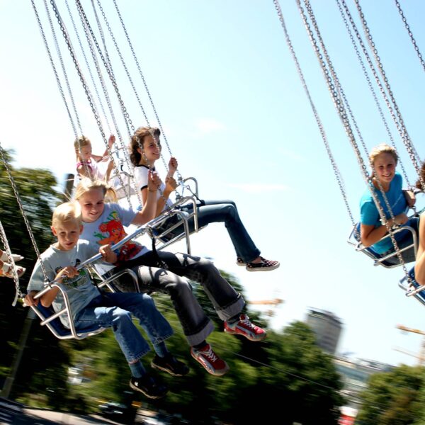 Kinder fahren auf einem sich drehenden Schaukelkarussell in einem Vergnügungspark unter einem klaren blauen Himmel, mit Bäumen und Gebäuden im Hintergrund.