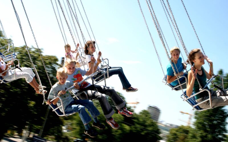 Kinder fahren auf einem sich drehenden Schaukelkarussell in einem Vergnügungspark unter einem klaren blauen Himmel, mit Bäumen und Gebäuden im Hintergrund.