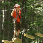 Ein Kind, das einen Helm und ein Geschirr trägt, läuft auf einem aufgehängten hölzernen Hindernisparcours in einem Wald.