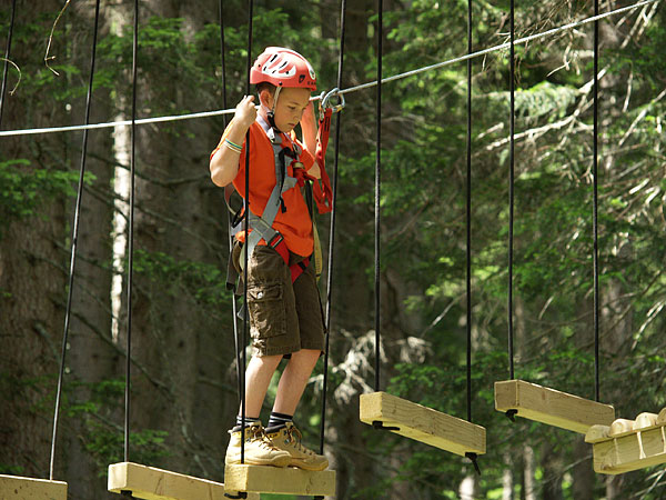 Ein Kind, das einen Helm und ein Geschirr trägt, läuft auf einem aufgehängten hölzernen Hindernisparcours in einem Wald.