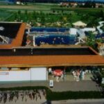 Luftaufnahme eines Freibadkomplexes mit schwimmenden und sonnenbadenden Menschen und Fahrrädern, die in der Nähe des Eingangs abgestellt sind.