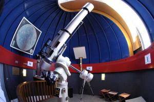 Ein großes Teleskop ist in einem kuppelförmigen Observatorium mit blauen Wänden und einer gewölbten Decke montiert. An den Wänden sind ein Bild des Mondes und Wegweiser zu sehen.