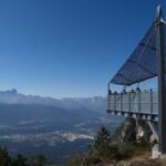 Eine erhöhte Aussichtsplattform mit einem schattenspendenden Vordach bietet einen Blick auf ein Tal und eine entfernte Bergkette unter einem klaren blauen Himmel.
