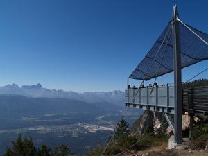 Eine erhöhte Aussichtsplattform mit einem schattenspendenden Vordach bietet einen Blick auf ein Tal und eine entfernte Bergkette unter einem klaren blauen Himmel.