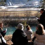 Eine Gruppe von Menschen sitzt im Freien auf einem Hügel in der Nähe eines Flusses und hört einer Person zu, die steht und spricht, wobei im Hintergrund eine Landschaft zu sehen ist.