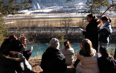 Eine Gruppe von Menschen sitzt im Freien auf einem Hügel in der Nähe eines Flusses und hört einer Person zu, die steht und spricht, wobei im Hintergrund eine Landschaft zu sehen ist.