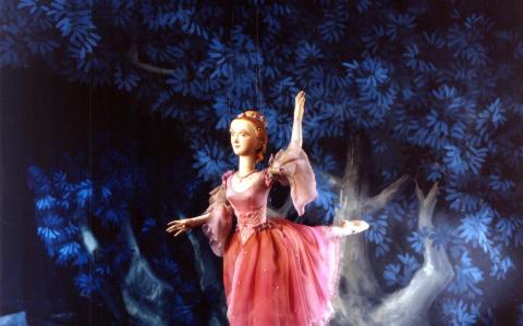 Eine Marionettenballerina in einem rosafarbenen Kostüm steht auf einem Bein vor einem blauen Blätterhintergrund mit nebelartigen Effekten zu ihren Füßen.