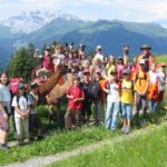Eine Gruppe von Kindern und Erwachsenen posiert mit Lamas auf einem grasbewachsenen Bergpfad, mit grünen Hügeln und schneebedeckten Gipfeln im Hintergrund.
