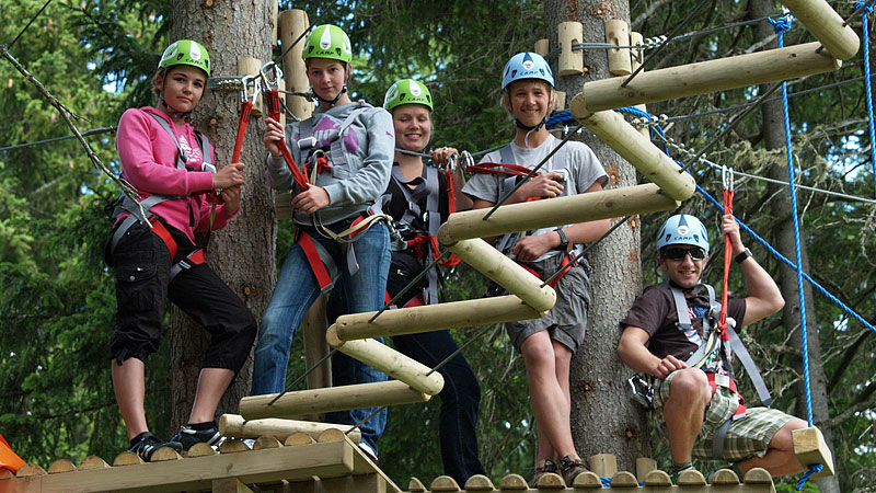 Fünf Personen mit Helmen und Gurten stehen auf einer hölzernen Plattform in einem Baumkronen-Parcours, umgeben von Bäumen und Seilhindernissen.
