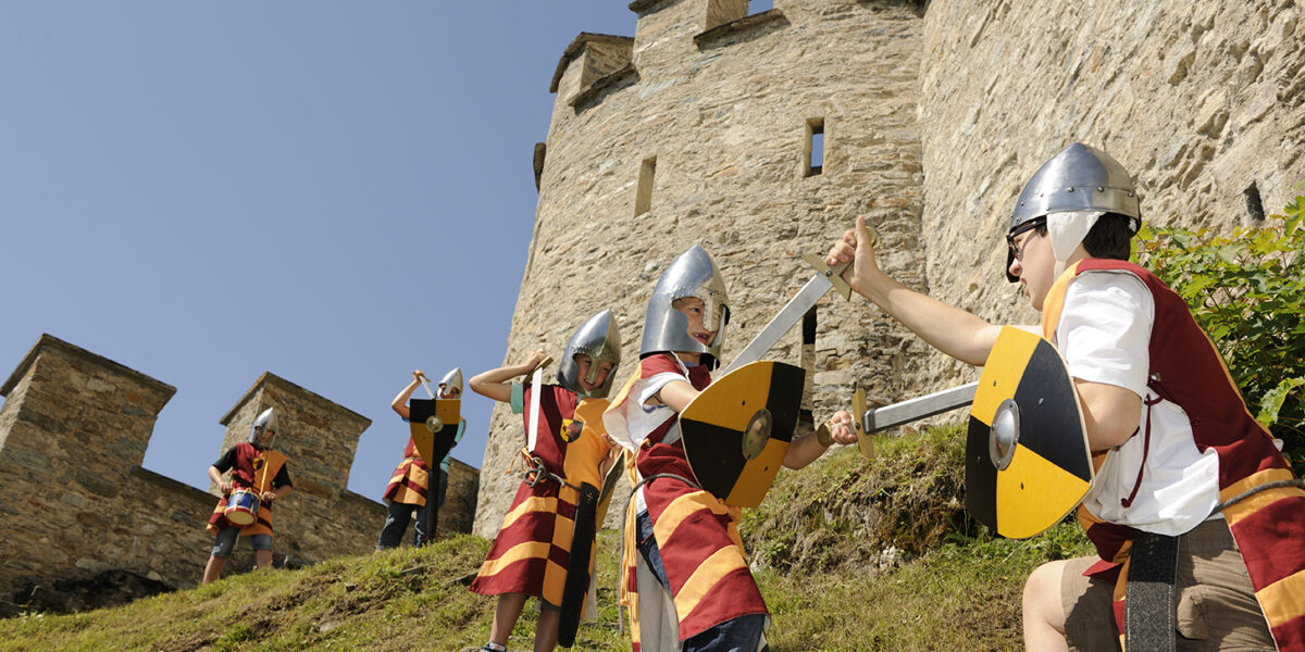Kinder mit Spielzeugrüstungen und -helmen stellen an einem sonnigen Tag vor einer Steinburg spielerisch eine mittelalterliche Schlacht mit Schilden und Schwertern nach.