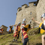 Kinder mit Spielzeugrüstungen und -helmen stellen an einem sonnigen Tag vor einer Steinburg spielerisch eine mittelalterliche Schlacht mit Schilden und Schwertern nach.